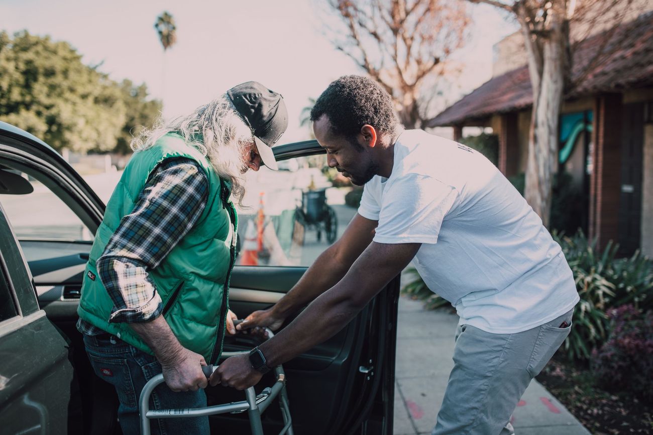A support worker helping an older man with a walking frame into a car